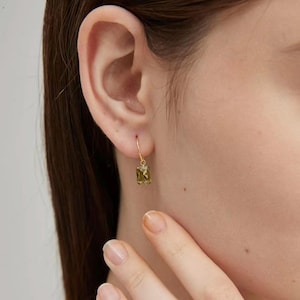 Puede incluir: Primer plano de un pendiente dorado con una gema rectangular verde oliva. El pendiente es de estilo colgante, con un fino aro dorado texturizado. La gema est&aacute; engastada en un bisel dorado y tiene una superficie facetada.