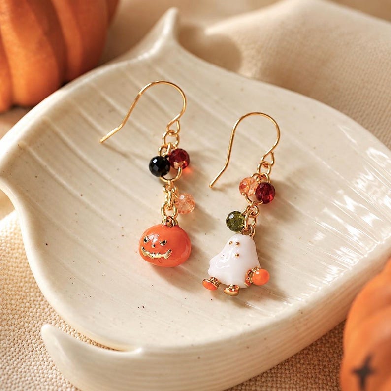 May include: A pair of gold-tone drop earrings featuring Halloween-themed charms. One earring displays an orange jack-o'-lantern, while the other features a white ghost. Both earrings are accented with small, multi-colored faceted beads.