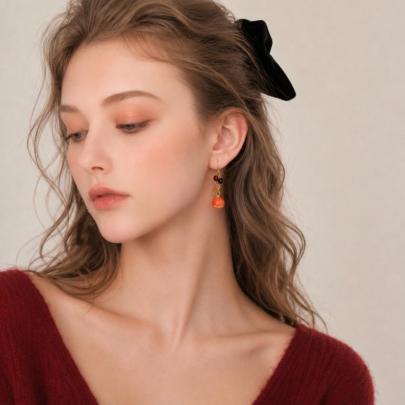 May include: A gold-toned drop earring featuring a small, bright orange pumpkin charm suspended from a delicate chain, accented with a tiny dark bead near the top hook, worn on an earlobe with a dark velvet hair bow visible in the hair.