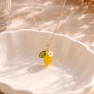 May include: A gold-colored necklace with a pendant featuring a yellow lemon, a green leaf, and a white flower. The lemon pendant has a textured surface. The necklace is displayed on a white, scalloped dish.