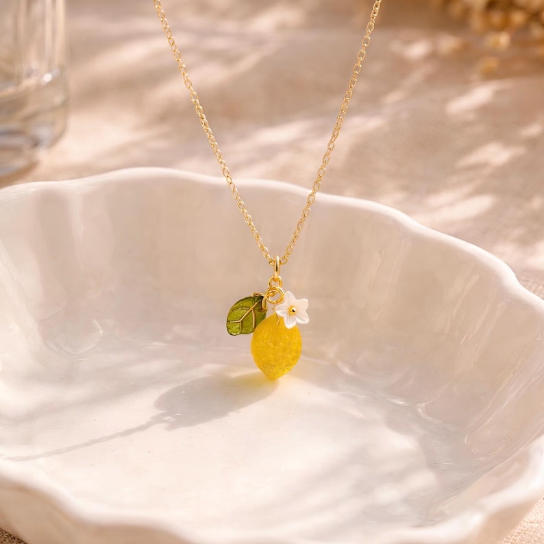 May include: A gold-colored necklace with a pendant featuring a yellow lemon, a green leaf, and a white flower. The lemon pendant has a textured surface. The necklace is displayed on a white, scalloped dish.