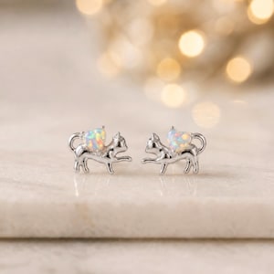 May include: A pair of silver cat-shaped stud earrings. Each earring features a cat silhouette with a heart-shaped opal stone. The opal stones display a mix of colors. The earrings are set against a blurred background.