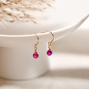 May include: A pair of gold-coloured drop earrings featuring bright pink, teardrop-shaped gemstones. The earrings have curved ear wires. The background is soft white, with a hint of dried grasses.