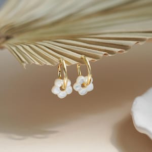 May include: Gold hoop earrings with white flower pendants. The earrings are set against a neutral backdrop with a dried palm leaf. These earrings are a delicate and feminine accessory, suitable for everyday wear.