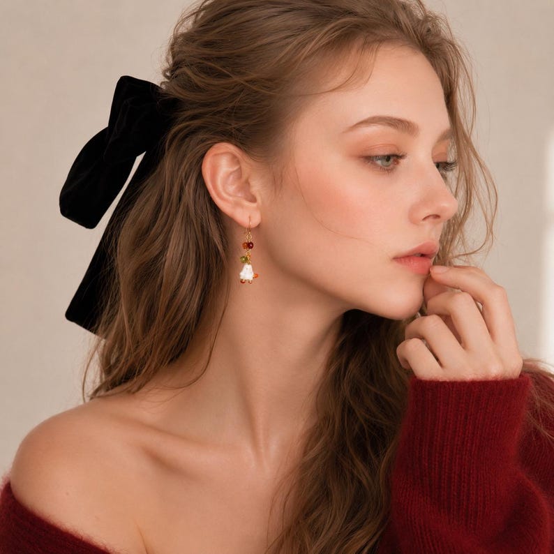 May include: A gold-toned drop earring featuring a small white flower-shaped charm, a tiny green bead, and a small red bead suspended from a thin gold wire hook. The earring is worn on the earlobe, showcasing a delicate and lightweight design.