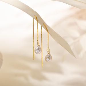 Könnte beinhalten: Ein Paar goldene Kettenohrringe mit jeweils einer einzelnen weißen Perle, die an der Kette baumelt.