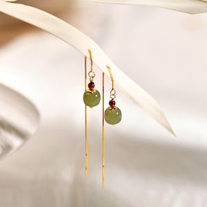 Może przedstawiać: Para złotych kolczyków wiszących z zielonymi koralikami z jadeitu i czerwonymi kamieniami szlachetnymi.