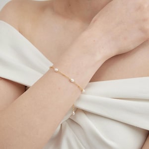 May include: A delicate gold bracelet featuring several oval-shaped pearls. The bracelet is worn on a wrist, with a simple design and a small dangling pearl charm. The background is a white fabric.