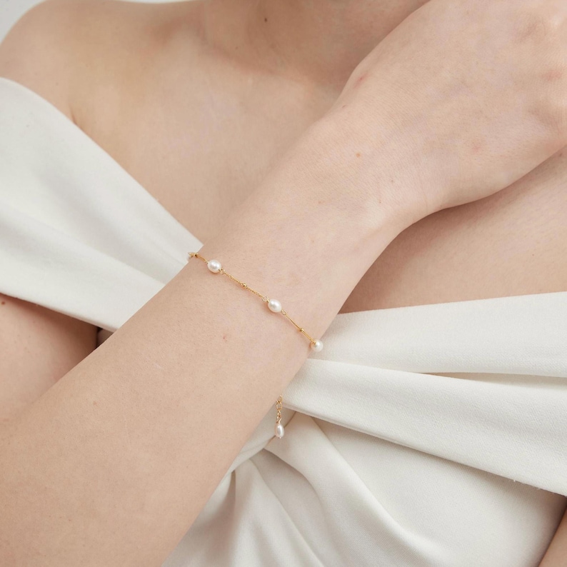 May include: A delicate gold bracelet featuring several oval-shaped pearls. The bracelet is worn on a wrist, with a simple design and a small dangling pearl charm. The background is a white fabric.
