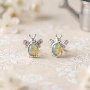 May include: A pair of silver bee-shaped stud earrings featuring oval-cut iridescent opals as the body. The wings are detailed with etched lines, and the head includes two small antennae. The metal has a polished finish, and the opals display flashes of orange, yellow, and green colours.