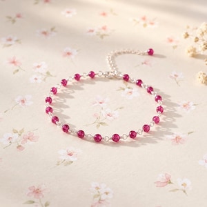May include: A delicate bracelet featuring a series of small, faceted, deep pink beads linked together by thin silver-toned metal wire. The bracelet includes a short extender chain at one end, finished with a single matching pink bead at the tip.