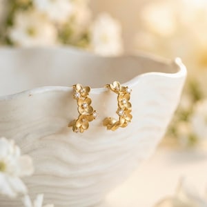 Könnte beinhalten: Ein Paar goldene Creolen, die mit einer Ansammlung kleiner, mattierter Blumenformen am Außenrand verziert sind. Jede Blume ist in der Mitte mit einem winzigen, klaren, funkelnden Edelstein akzentuiert, was ein zartes, florales Design ergibt.