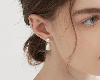 May include: A drop earring featuring a small, round white pearl stud connected to a larger, teardrop-shaped white pearl pendant. The earring is crafted with a simple metal post and backing, showcasing a clean, polished aesthetic with smooth surfaces.