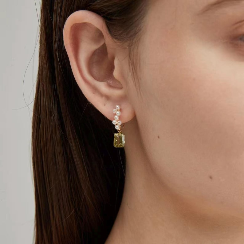 Puede incluir: Pendientes dorados con un grupo de peque&ntilde;as perlas y una gema rectangular verde oliva. Los pendientes son colgantes y tienen un dise&ntilde;o delicado, mostrando una mezcla de elementos cl&aacute;sicos y modernos. Los pendientes se llevan en una oreja.