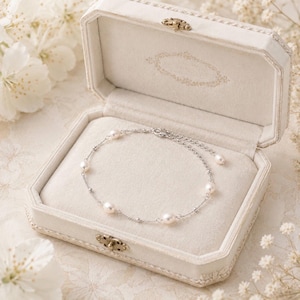 May include: A delicate pearl bracelet rests inside an open, white jewelry box. The bracelet features several round pearls strung on a silver chain. The box is octagonal with a decorative clasp and a soft, cream-colored interior.
