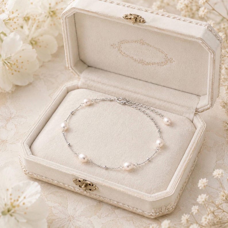 May include: A delicate pearl bracelet rests inside an open, white jewelry box. The bracelet features several round pearls strung on a silver chain. The box is octagonal with a decorative clasp and a soft, cream-colored interior.