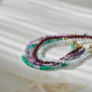 Pu&ograve; includere: Una collezione di delicati braccialetti di perline in vari colori, tra cui verde, viola, rosso e blu. I braccialetti sono fatti di piccole perline rotonde e sono disposti a strati. Lo sfondo &egrave; bianco con ombre morbide.