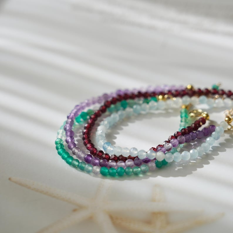 Pu&ograve; includere: Una collezione di delicati braccialetti di perline in vari colori, tra cui verde, viola, rosso e blu. I braccialetti sono fatti di piccole perline rotonde e sono disposti a strati. Lo sfondo &egrave; bianco con ombre morbide.