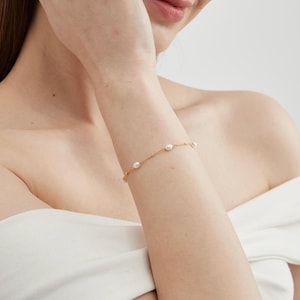 May include: A delicate gold bracelet featuring small, round pearls evenly spaced along a thin chain. The bracelet is worn on a wrist, with the skin tone appearing fair. The background is a soft, neutral color, highlighting the jewelry.