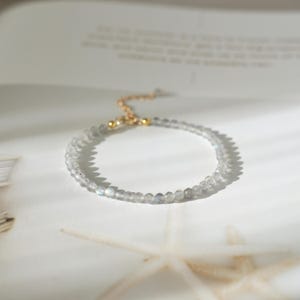 Op de afbeelding: Een delicate armband met kleine, ronde, lichtgekleurde kralen. De armband heeft een goudkleurige sluiting en ketting. De kralen lijken semi-transparant en de armband ligt op een wit oppervlak.