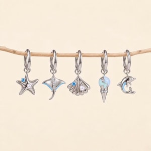 Peut inclure: Cinq boucles d'oreilles créoles argentées avec des breloques pendantes : une étoile de mer avec une pierre bleue et une perle, une raie avec des accents bleus, un coquillage avec une pierre bleue et une perle, une glace et un dauphin bleu.