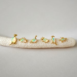 May include: A collection of six gold-colored dinosaur-shaped earrings with iridescent opal-like centers. The earrings are arranged in a row on a white, textured surface. The dinosaurs vary in design, showcasing different species.