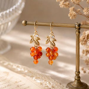 Peut inclure: Une paire de boucles d'oreilles pendantes dorées composées de grappes de perles rondes et translucides orange en forme de raisin. Chaque boucle présente une petite feuille en métal doré, un cristal transparent et un crochet d'oreille doré.