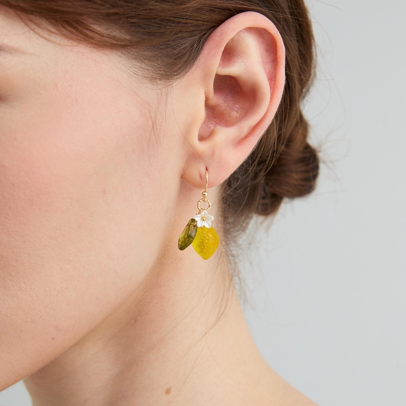 May include: Gold-tone drop earrings featuring a yellow lemon and green leaf design. The earrings have a small white flower accent. The earrings are shown on a person's ear.