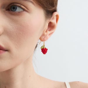 May include: A close-up of a gold-colored earring with a red raspberry design. The raspberry is made of small, round red beads. A small green leaf is attached to the top of the raspberry.