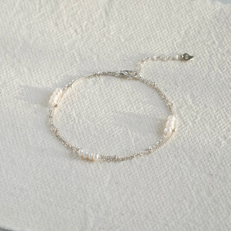 May include: A delicate silver chain bracelet with small white pearls. The bracelet has a double chain design and a heart-shaped clasp.