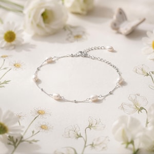 May include: A delicate silver bracelet featuring oval-shaped pearls. The bracelet is displayed on a white surface with floral accents, including daisies and other white flowers. The bracelet has a simple, elegant design.