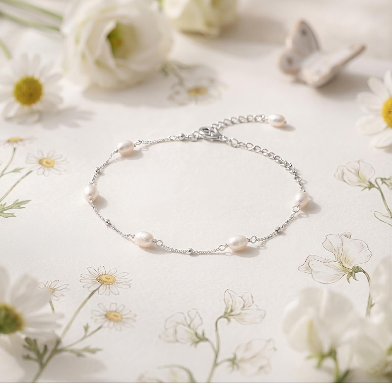 May include: A delicate silver bracelet featuring oval-shaped pearls. The bracelet is displayed on a white surface with floral accents, including daisies and other white flowers. The bracelet has a simple, elegant design.