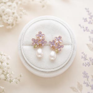 May include: A pair of floral earrings with a light purple flower design and a dangling white pearl. The earrings are set on a gold-colored metal and are displayed in a light blue velvet jewelry box.