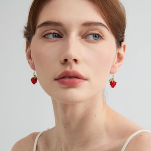 May include: A pair of red raspberry earrings with gold-colored hooks. Each earring features a cluster of red beads, with a small green leaf accent. The earrings are designed to dangle and add a touch of whimsy.