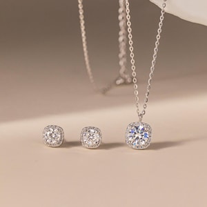 May include: A silver-toned jewelry set consisting of a necklace and two stud earrings. Each piece features a central round brilliant-cut stone surrounded by a halo of smaller pavé stones. The necklace includes a delicate silver-toned chain.