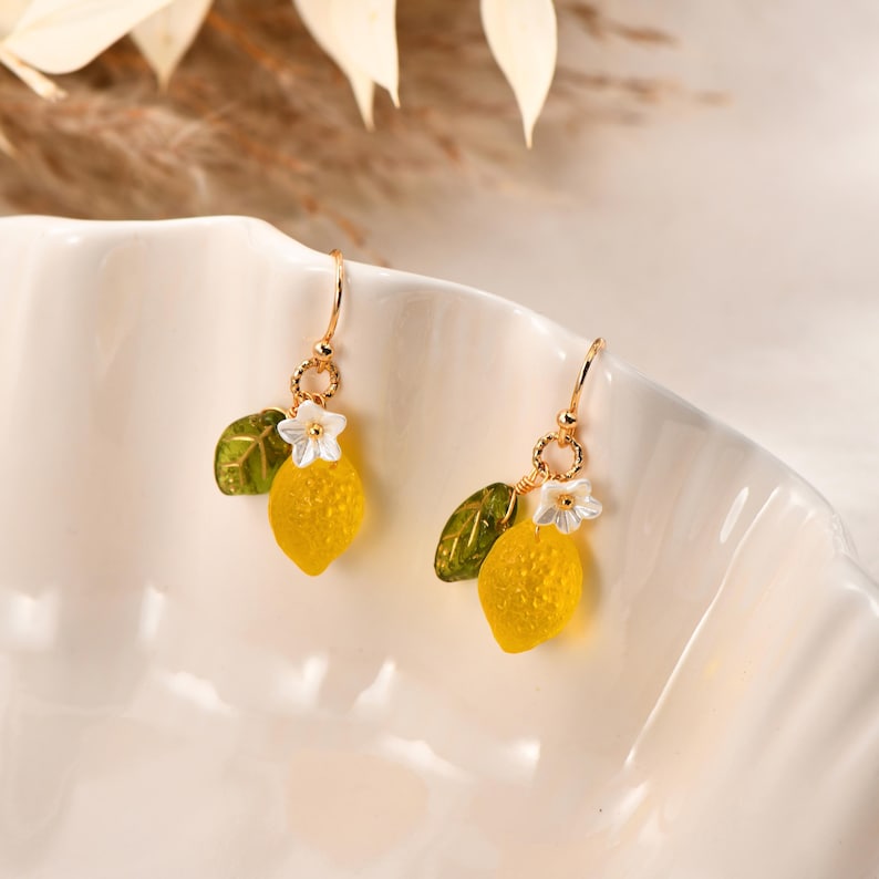 May include: A pair of gold-toned drop earrings featuring yellow lemon charms with green leaves and white flower accents. The earrings are suspended from delicate gold hooks, creating a summery, fruit-themed accessory.