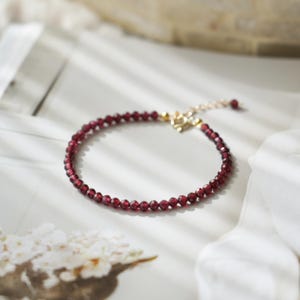 Könnte beinhalten: Ein zartes Armband mit kleinen, facettierten, tiefroten Perlen. Das Armband hat einen goldfarbenen Verschluss mit herzförmigem Design. Die Perlen sind aufgefädelt und bilden ein schlichtes, aber elegantes Schmuckstück.