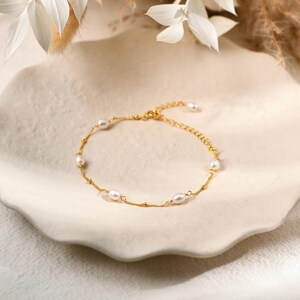 May include: A gold-colored bracelet with several white oval pearls and small gold beads. The bracelet is displayed on a white, textured, ceramic plate. The background includes white flowers and dried plants.