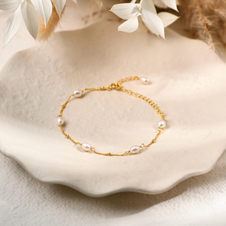 May include: A gold-colored bracelet with several white oval pearls and small gold beads. The bracelet is displayed on a white, textured, ceramic plate. The background includes white flowers and dried plants.