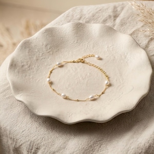May include: A delicate gold-colored bracelet with several white pearls. The bracelet is displayed on a white, scalloped-edge plate. The background is a neutral beige.