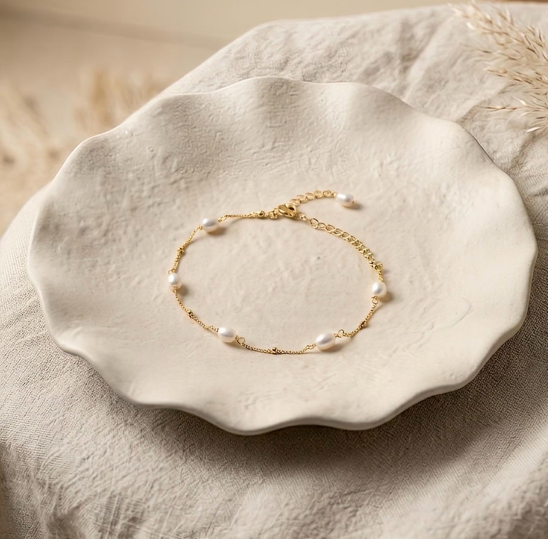May include: A delicate gold-colored bracelet with several white pearls. The bracelet is displayed on a white, scalloped-edge plate. The background is a neutral beige.