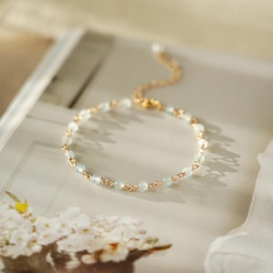 May include: A gold-coloured bracelet with small, pale blue beads. The bracelet has a delicate chain and clasp. The beads are evenly spaced along the chain. The bracelet is displayed on a light-coloured surface, with soft lighting.