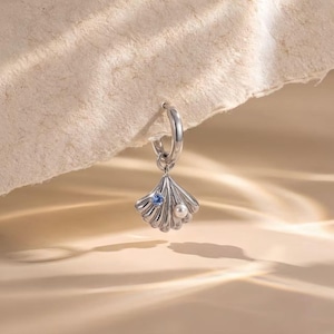 May include: A silver hoop earring featuring a dangling seashell charm. The shell is crafted from polished silver and is accented with a single small round blue gemstone and a small white pearl bead set into the textured surface of the metal.