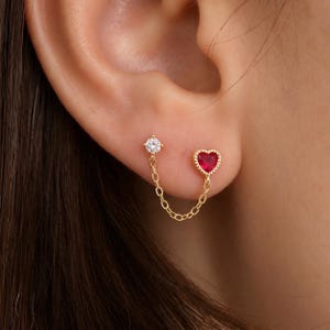 Puede incluir: Un pendiente doble de oro que presenta un perno con piedra redonda transparente y otro con piedra roja en forma de corazón, unidos por una delicada cadena dorada. La pieza de joyería está diseñada para llevarse en dos perforaciones adyacentes.