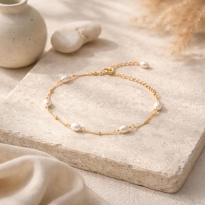 May include: A delicate gold-toned anklet featuring oval-shaped pearls. The anklet has a fine chain with small, round gold beads interspersed between the pearls. The clasp is visible, and the anklet is displayed on a neutral-toned surface.