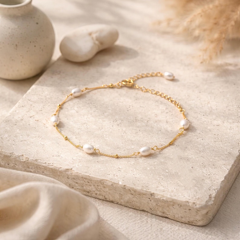May include: A delicate gold-toned anklet featuring oval-shaped pearls. The anklet has a fine chain with small, round gold beads interspersed between the pearls. The clasp is visible, and the anklet is displayed on a neutral-toned surface.