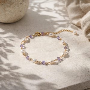 May include: A delicate gold bracelet featuring alternating freshwater pearls and small purple gemstones. The bracelet has a delicate chain and a secure clasp. The jewelry is displayed on a neutral surface.