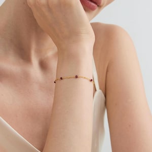 Op de afbeelding: Een delicate gouden armband met kleine, ronde, rode edelstenen. De armband wordt om een pols gedragen en toont de fijne ketting en de gelijkmatig verdeelde edelstenen. De sieraden zijn eenvoudig en elegant.