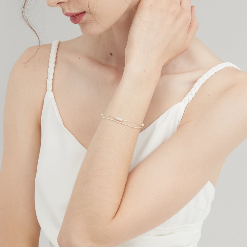 May include: A delicate silver bracelet with a row of small white pearls. The bracelet is layered with a thinner silver chain.