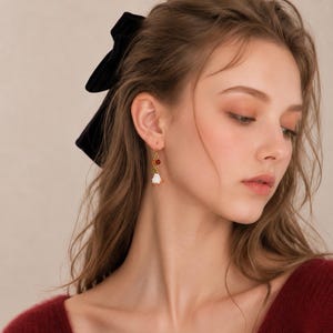 May include: A gold-toned dangle earring featuring a small white ghost charm with orange accents, suspended from a thin gold wire hook. The ghost charm is accented by two small, round, translucent beads in amber and green tones.